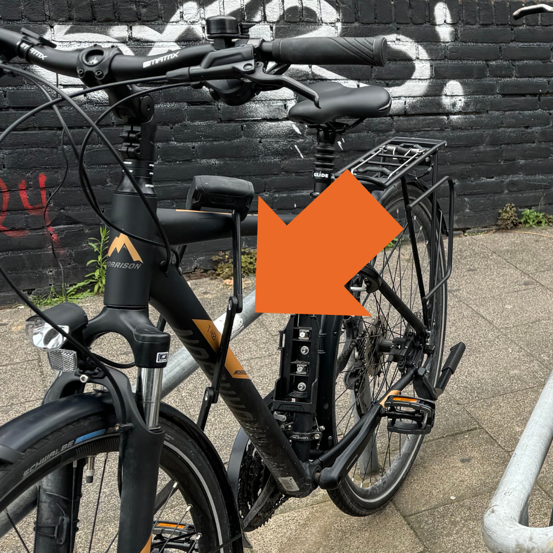 Fahrrad an einem eisernen Fahrradständer, auf dem ein Aufkleber klebt. Ein großer orangefarbener Pfeil weist auf den Aufkleber hin.